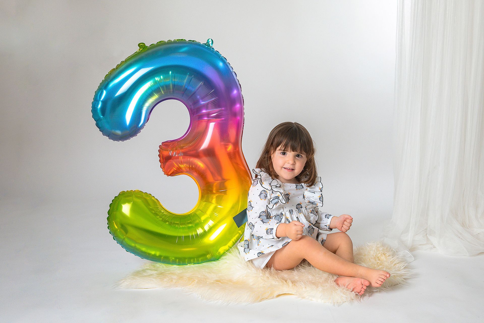 Una niña está sentada al lado de un globo con el número tres de color arcoíris.