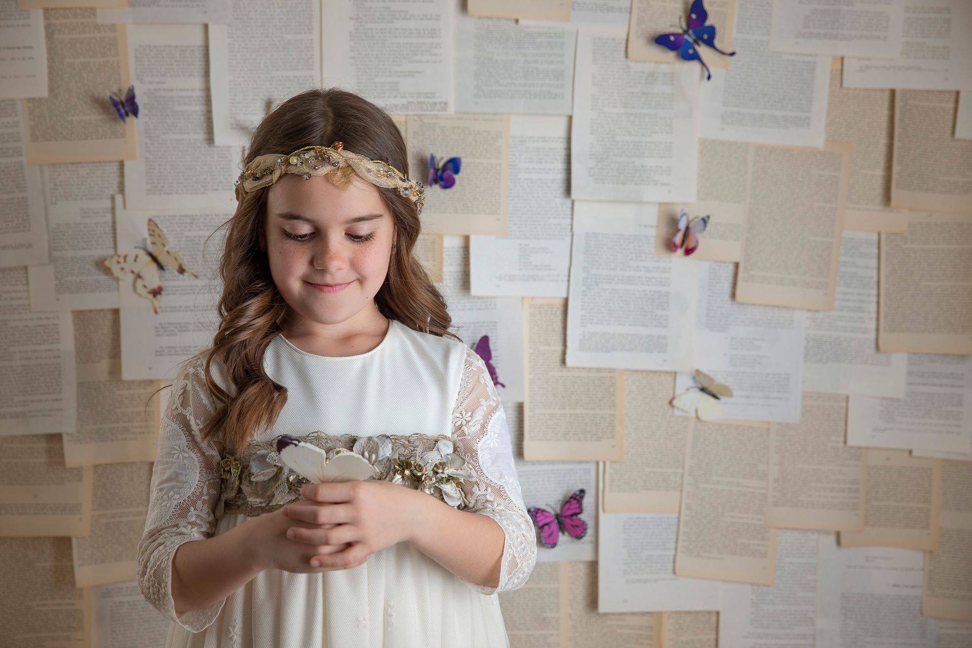 Una niña con un vestido blanco está parada frente a una pared de libros y mariposas.