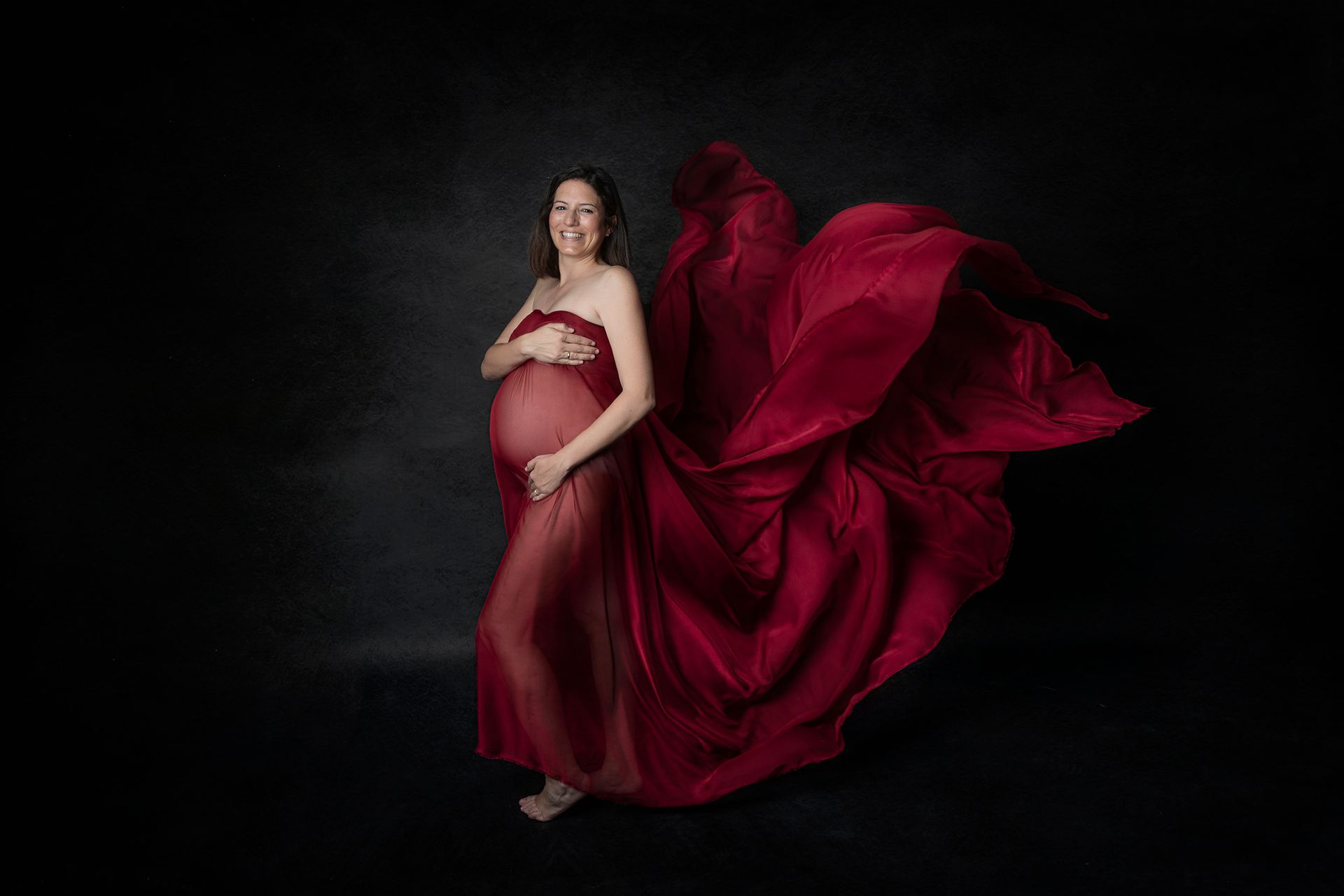 Una mujer embarazada con un vestido rojo está parada frente a un fondo negro.