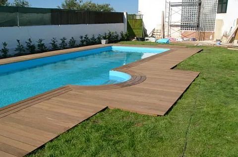 Piscina en el patio trasero con terraza de madera junto a un césped verde y agua azul.