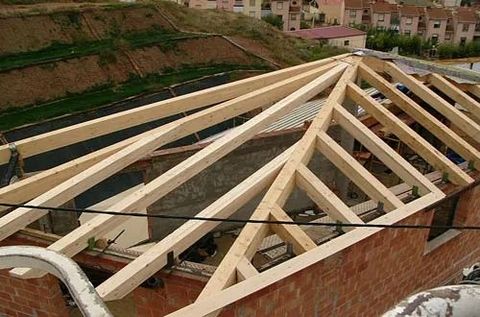 Cerchas de madera instaladas sobre un edificio de ladrillo en construcción, con una ladera y casas al fondo.