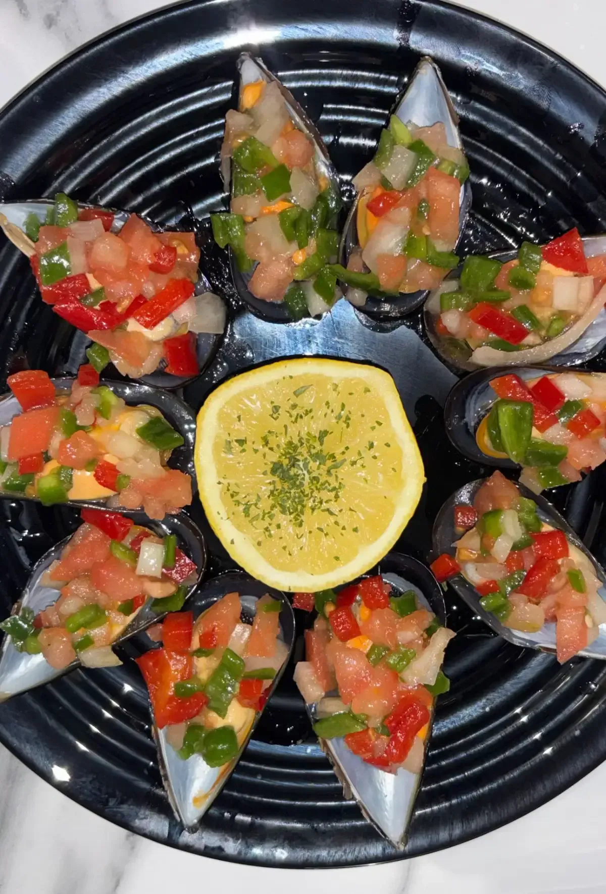 Una bandeja circular negra contiene diez mejillones cubiertos con una colorida salsa de tomate, cebolla y pimiento
