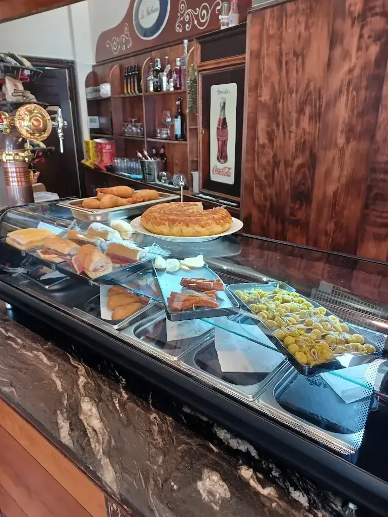 Un mostrador de cristal en una taberna que ofrece diversos aperitivos, una tortilla española y sándwiches en platos.