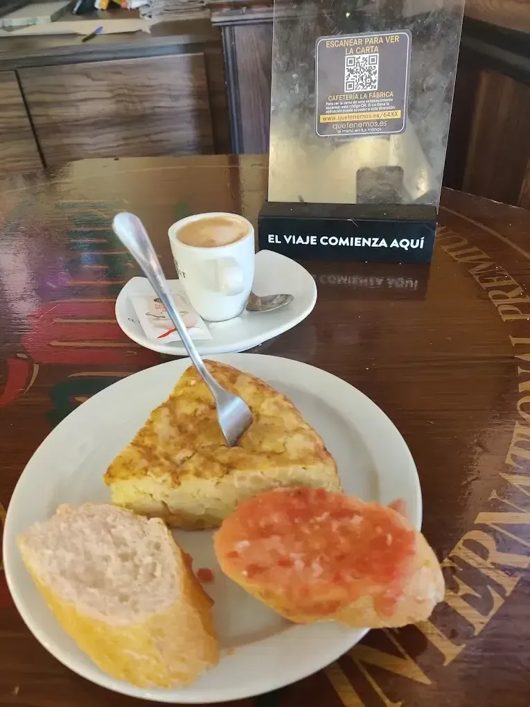 Un desayuno de tortilla, pan con tomate y un pequeño café servido en una mesa de madera con un cartel con código QR.