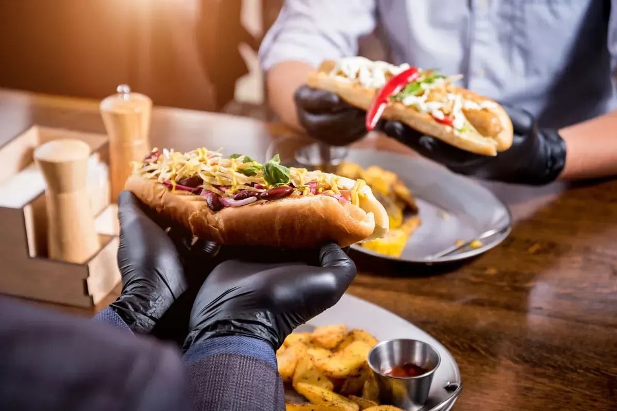 Una persona que lleva guantes negros sostiene un perrito caliente delante de un plato de patatas fritas.