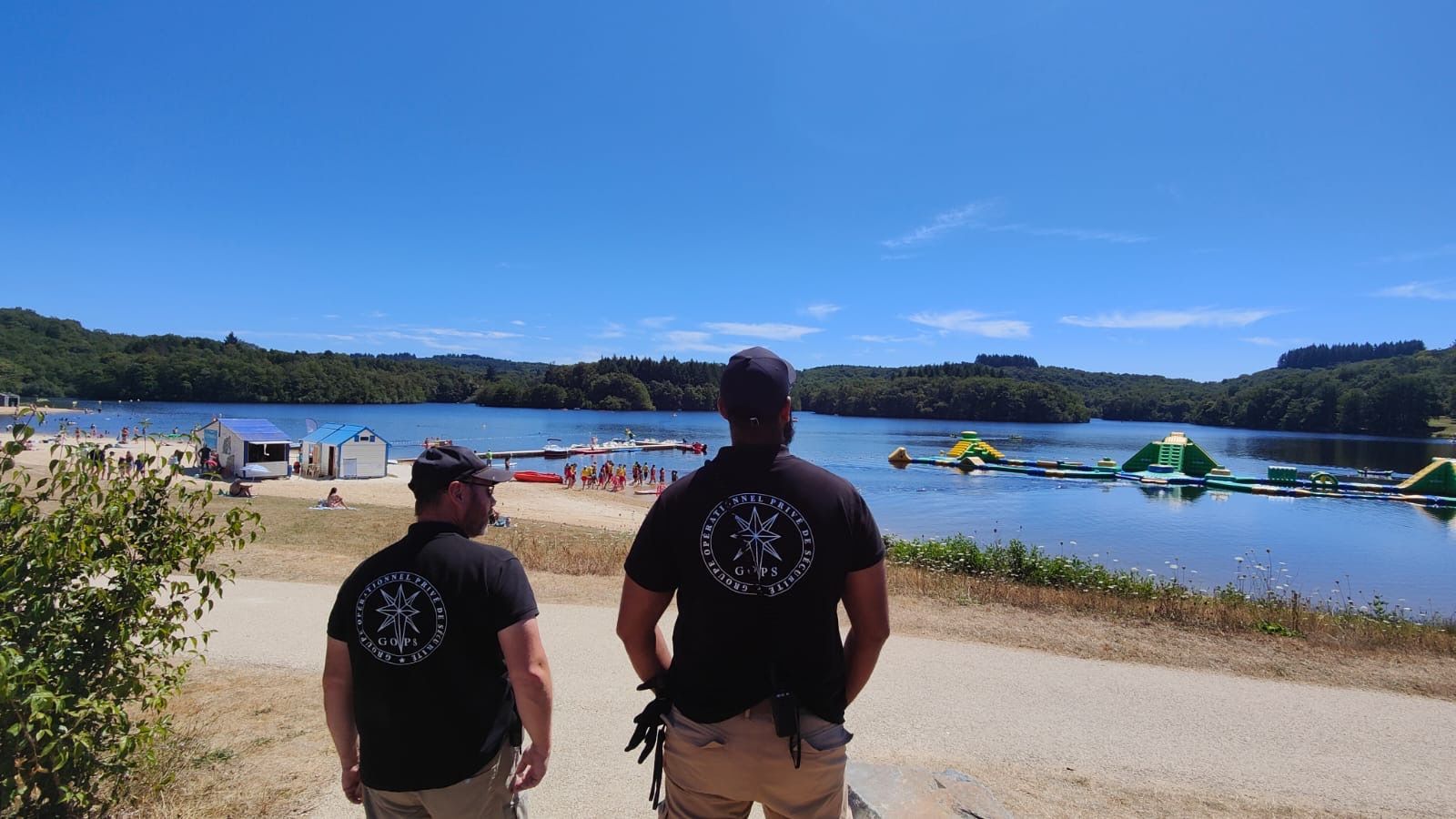 Deux hommes en chemises noires avec un logo, surplombant un lac avec une plage de sable et un parc aquatique gonflable par une journée ensoleillée.