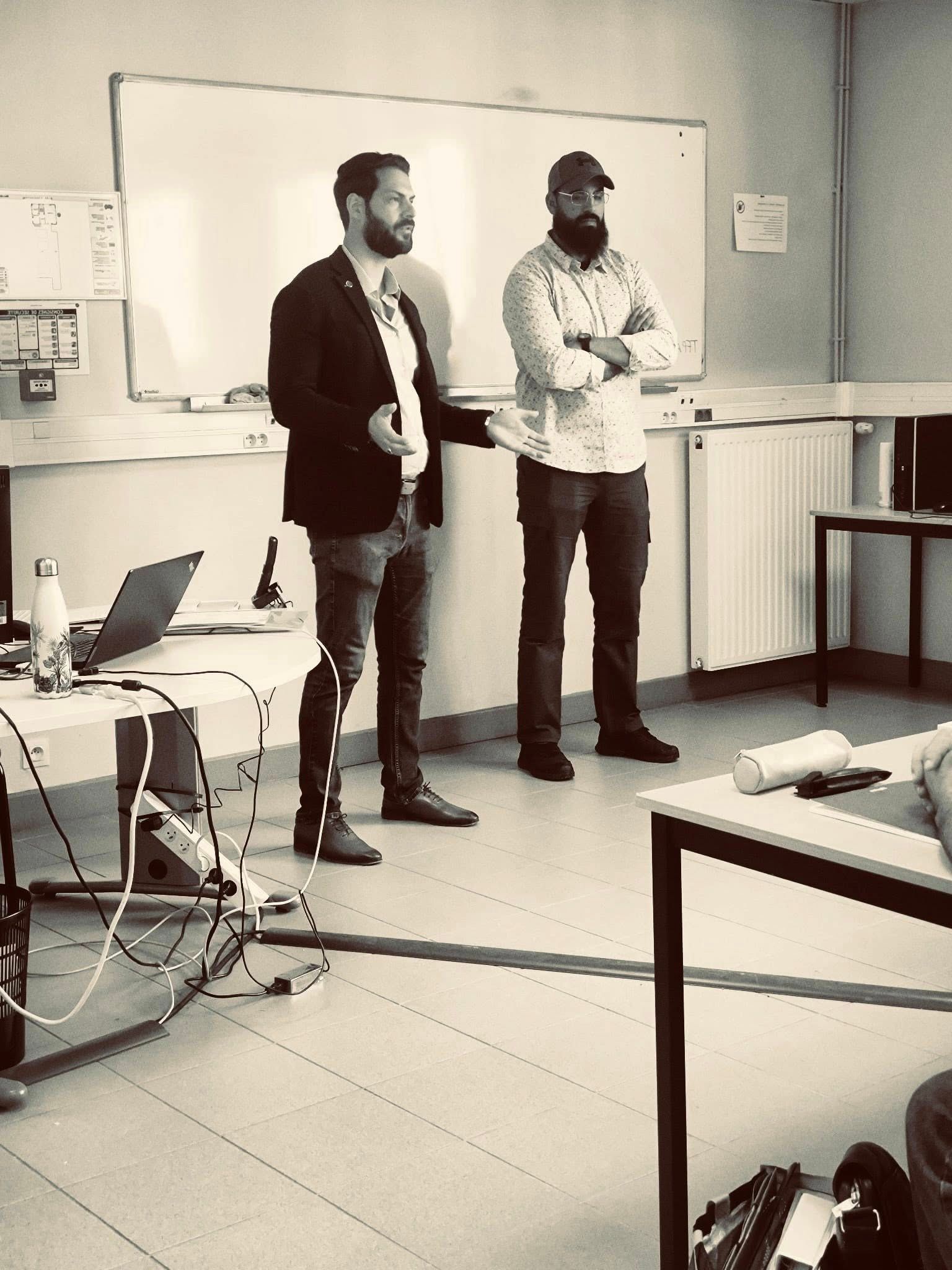 Deux hommes dans une salle de classe ; l'un, avec une prothèse de jambe, présente un exposé près d'un tableau blanc. L'autre, les bras croisés.