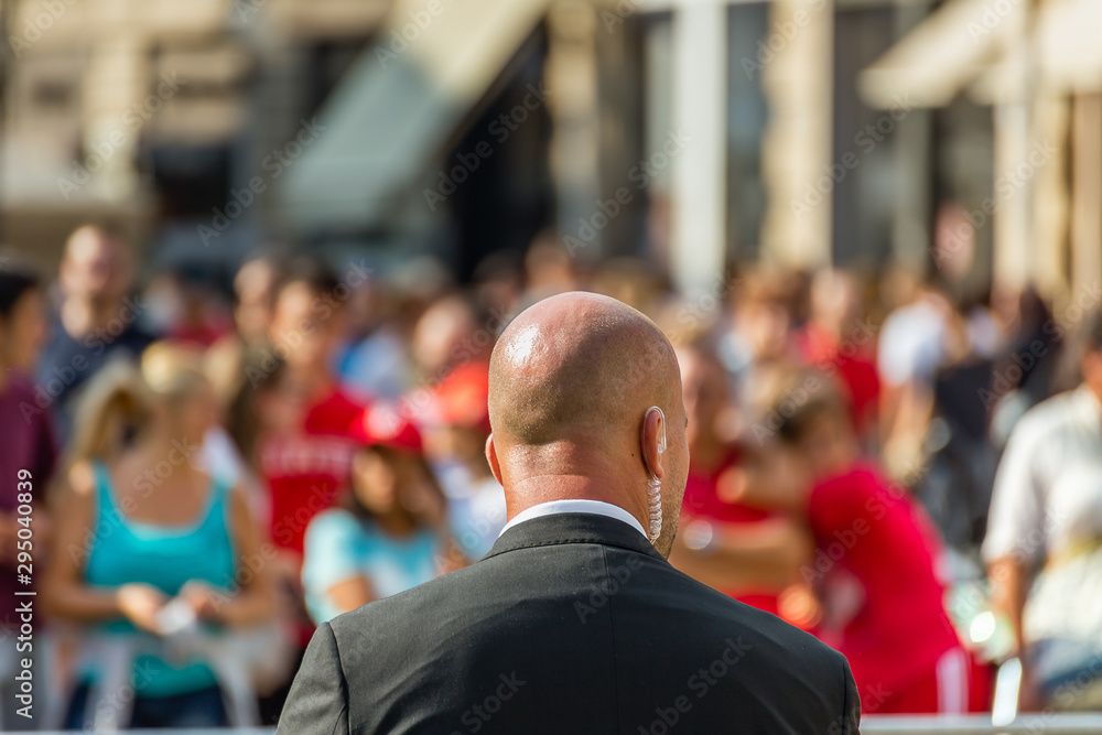 Homme chauve en costume, dos à la caméra, avec un écouteur, face à la foule de personnes à l'extérieur.