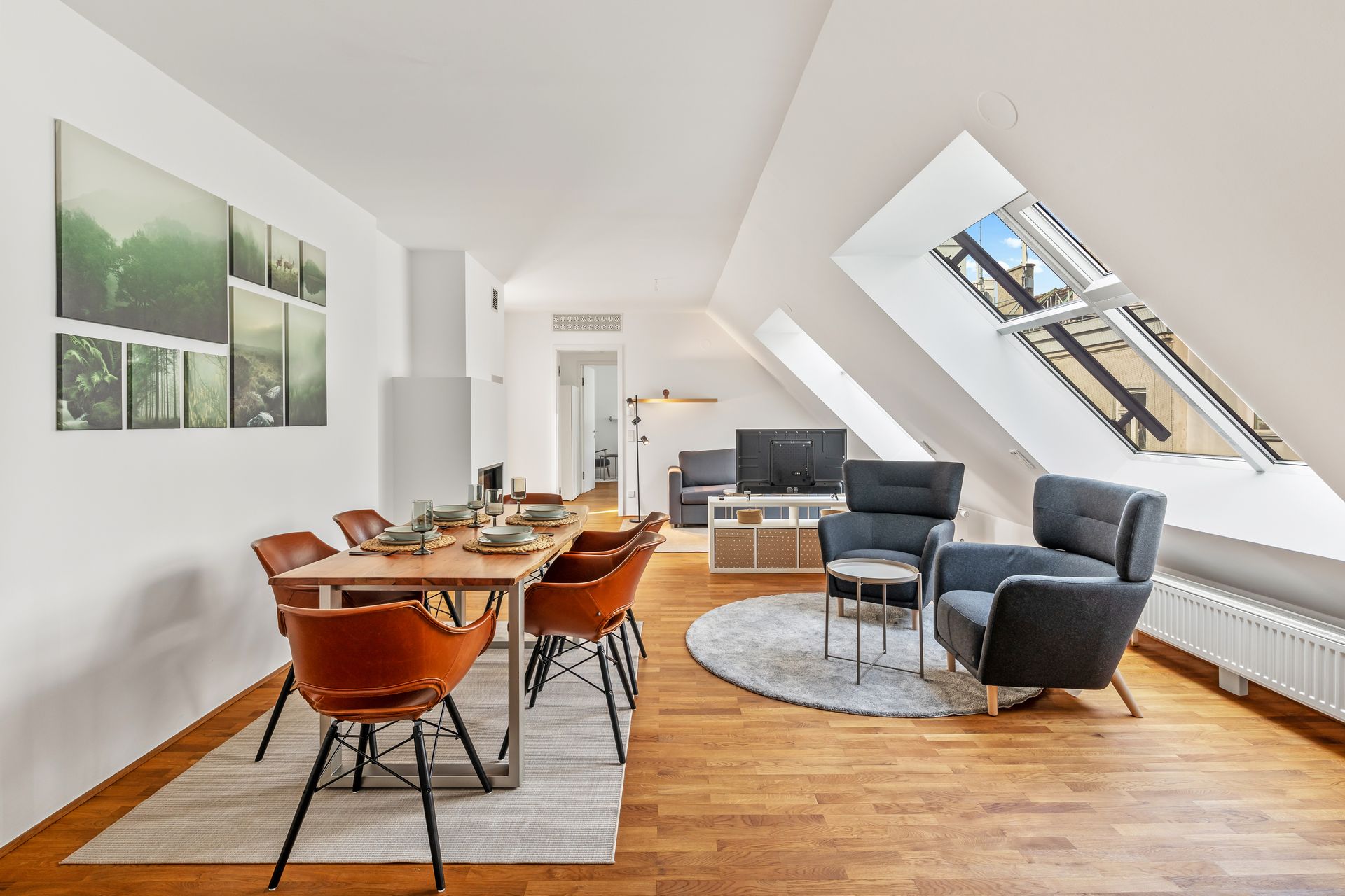 A bright, modern apartment interior. Dining table with orange chairs, living area with armchairs, and skylight.