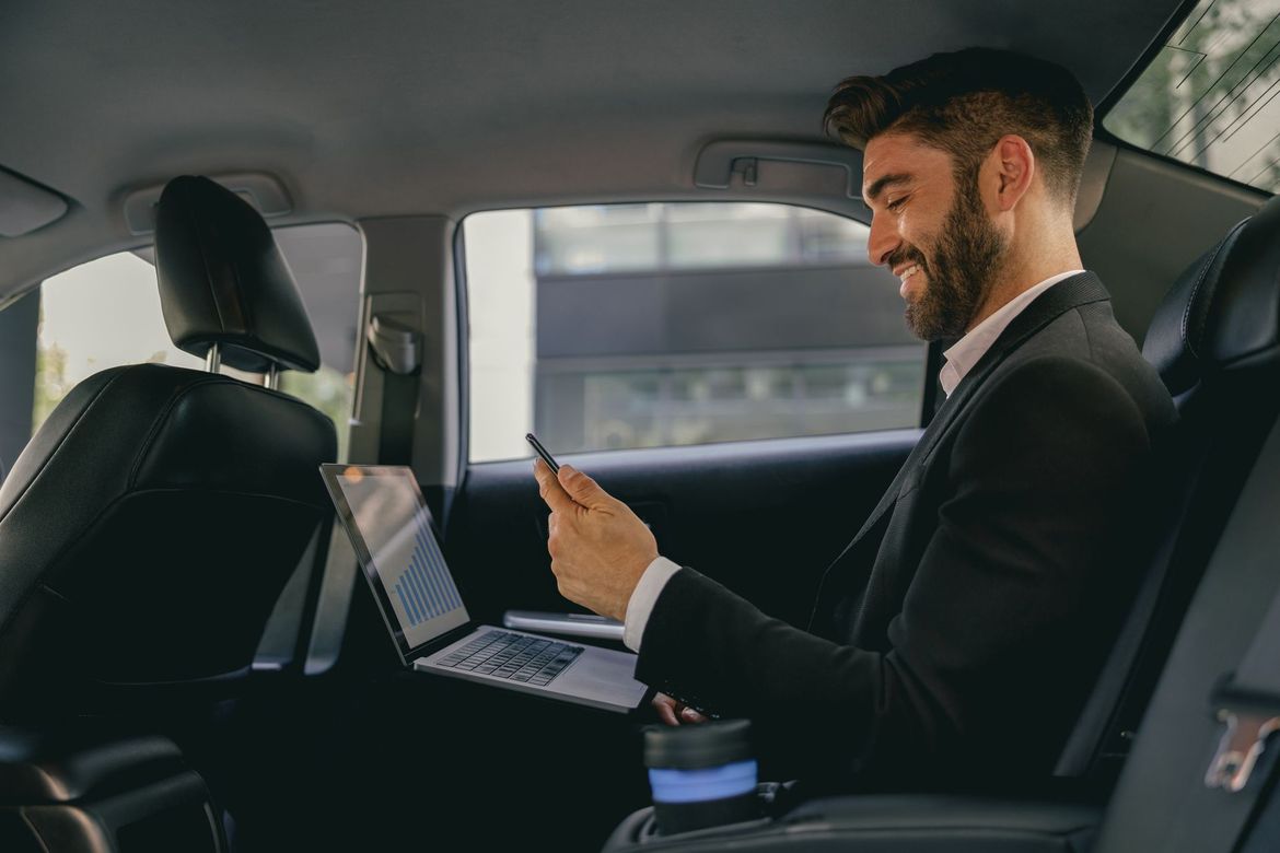Un homme en costume sourit et utilise son téléphone et son ordinateur portable sur la banquette arrière d'une voiture.