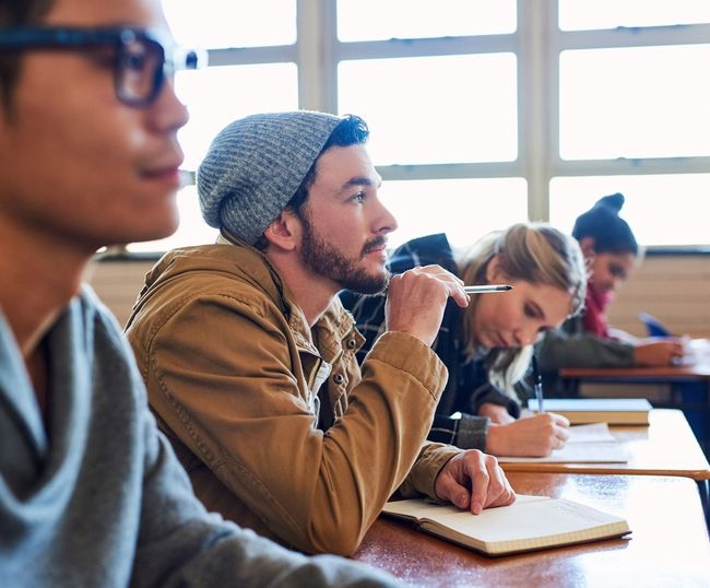 Un grupo de estudiantes están sentados en escritorios en un salón de clases.
