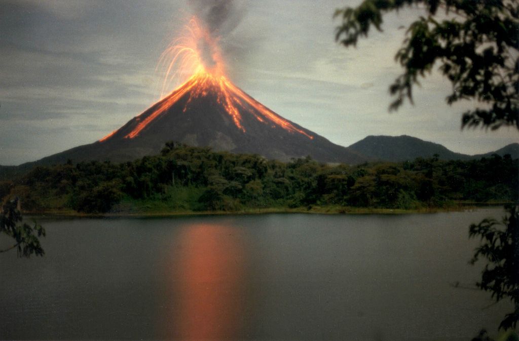 Arenal volcano is erupting over a lake with trees in the foreground