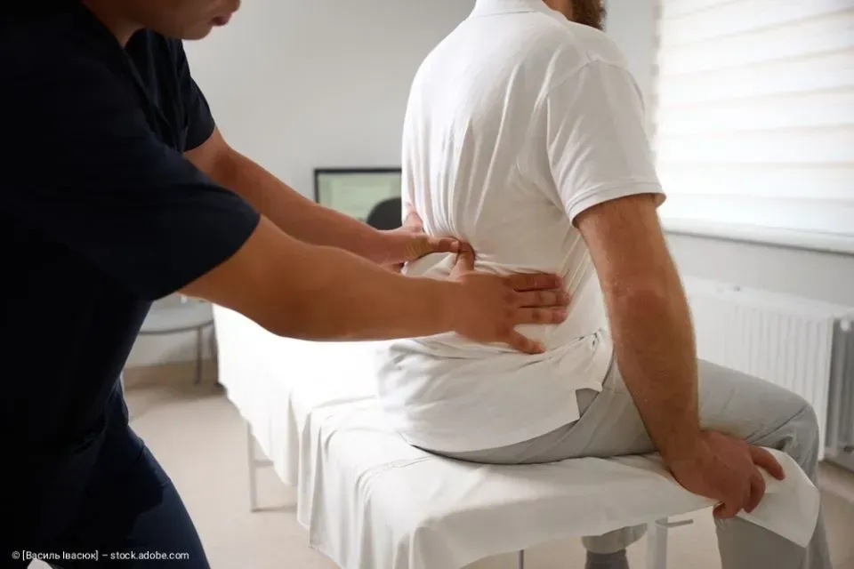 Eine Therapeutin massiert den Rücken eines sitzenden Patienten in einem hellen Behandlungsraum.