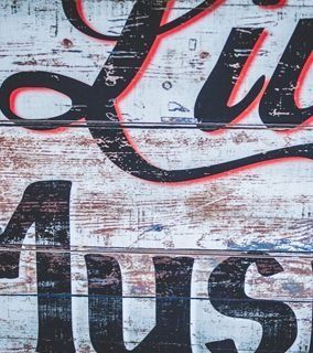 Letras negras sobre una superficie de madera desgastada y pintada de blanco, con líneas de paneles horizontales y detalles en rojo descolorido.