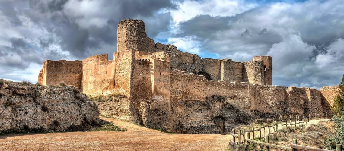 Las ruinas de los muros de piedra y la torre de un castillo histórico se alzan bajo un cielo nublado y dramático.