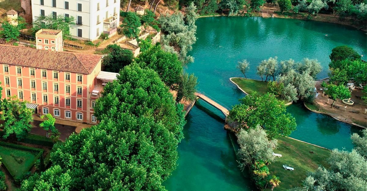 Un edificio de terracota de varias plantas situado junto a un río de un verde intenso, conectado por un estrecho puente de piedra a una orilla cubierta de hierba.