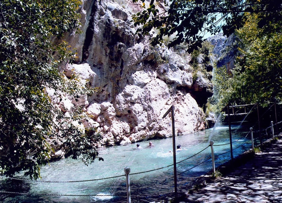 La gente nada en un arroyo de aguas cristalinas de color turquesa al pie de un gran acantilado de piedra caliza escarpada, rodeado de frondosos árboles.