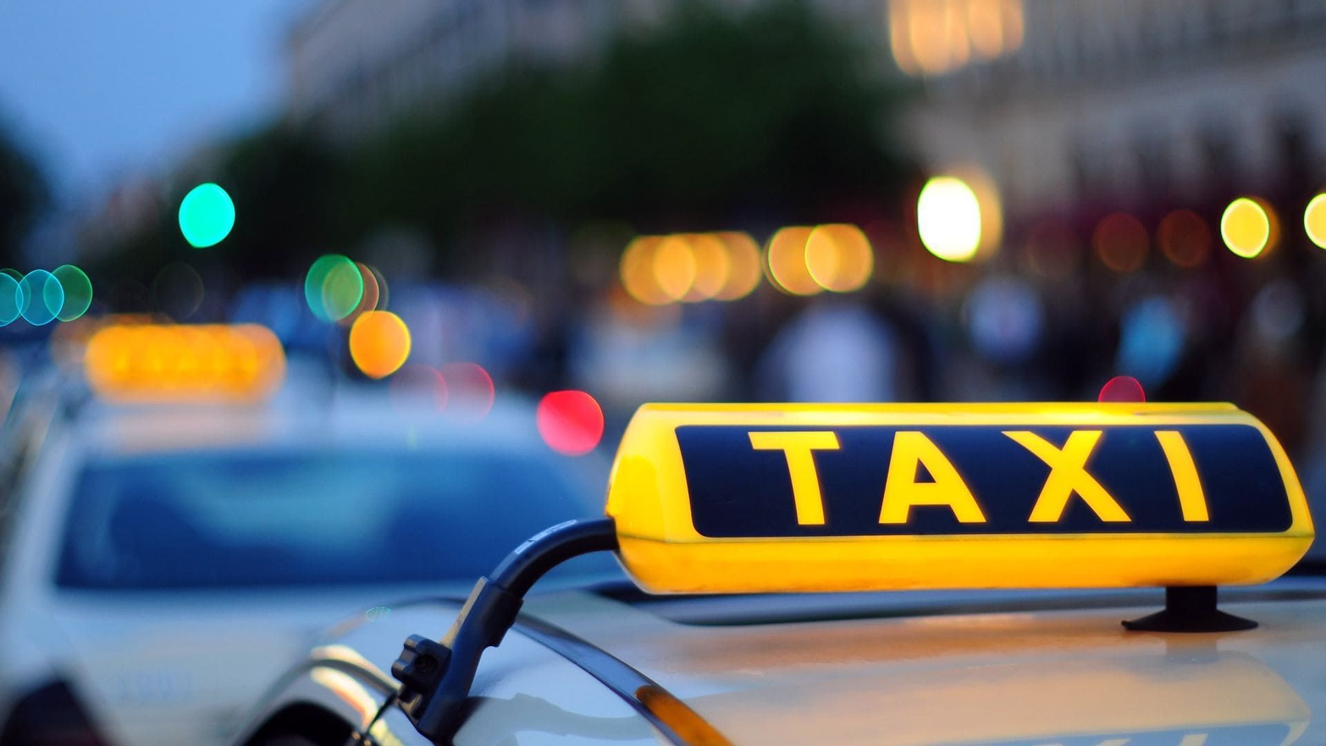 Una luz amarilla de taxi en el techo de un coche, con farolas borrosas al fondo, al anochecer.