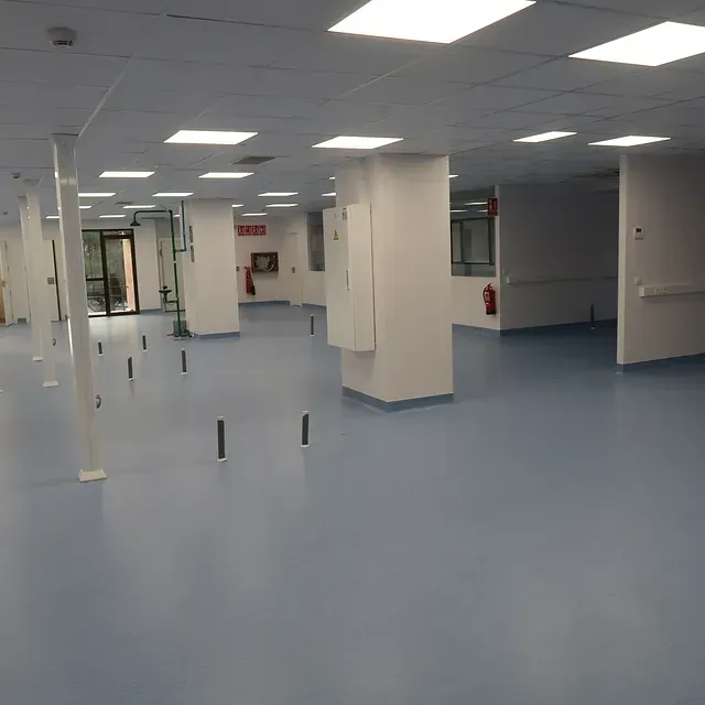 Habitación de hospital vacía, con suelo de color azul claro, columnas blancas, luces de techo y equipo de emergencia.