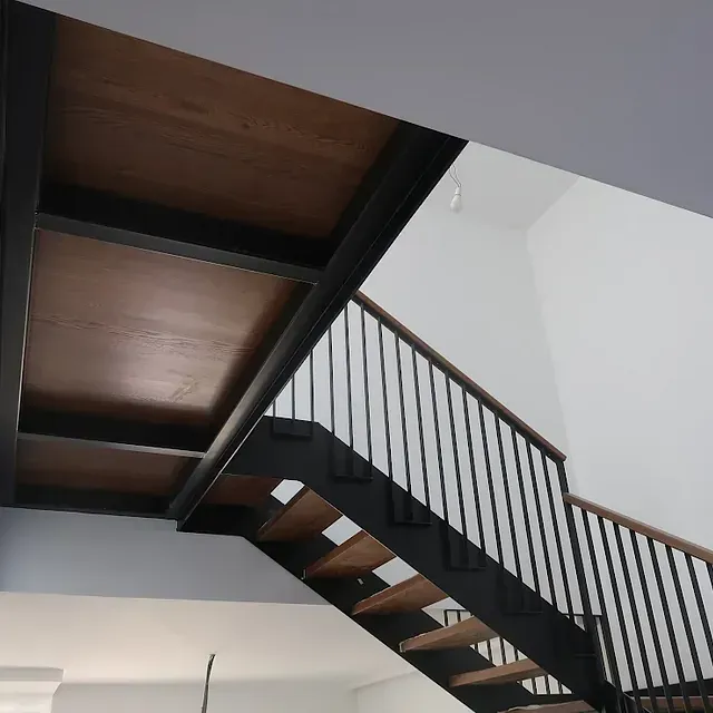 Escalera con peldaños de madera y barandilla de metal negro, bajo techo de madera y paredes blancas.