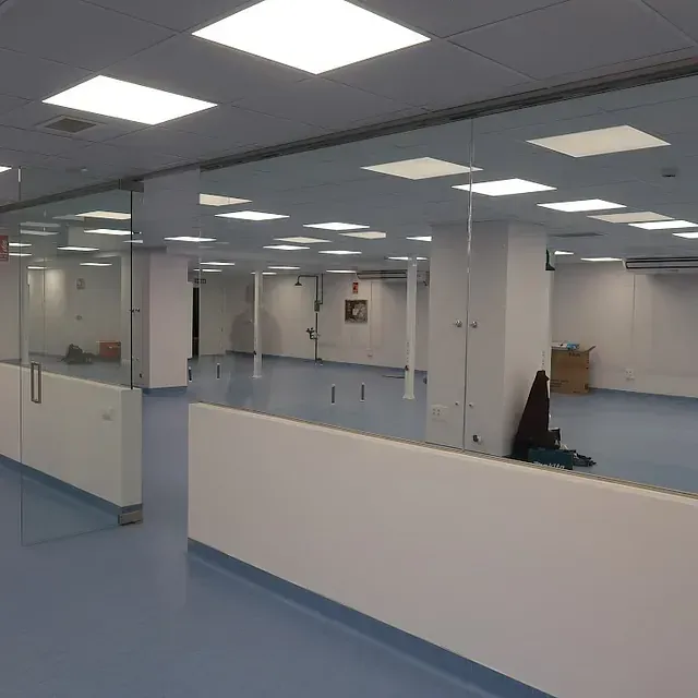 Habitación vacía y abierta con suelo azul, paredes y columnas blancas, luces de techo cuadradas y mamparas de cristal.