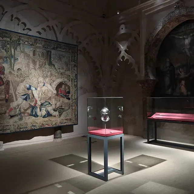 Exhibición de museo con tapiz, vitrinas y arquitectura arqueada.