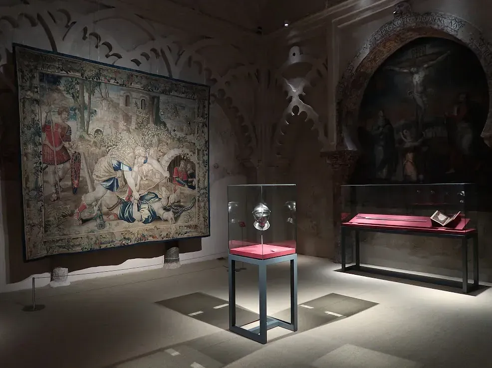 Exhibición del museo: Tapiz y objeto encapsulado con pintura en la pared en una sala con arquitectura gótica.