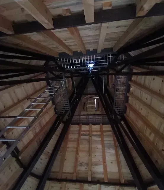 Dentro de un techo de cenador de madera, con una escalera, vigas de metal y una rejilla de ventilación en el centro.