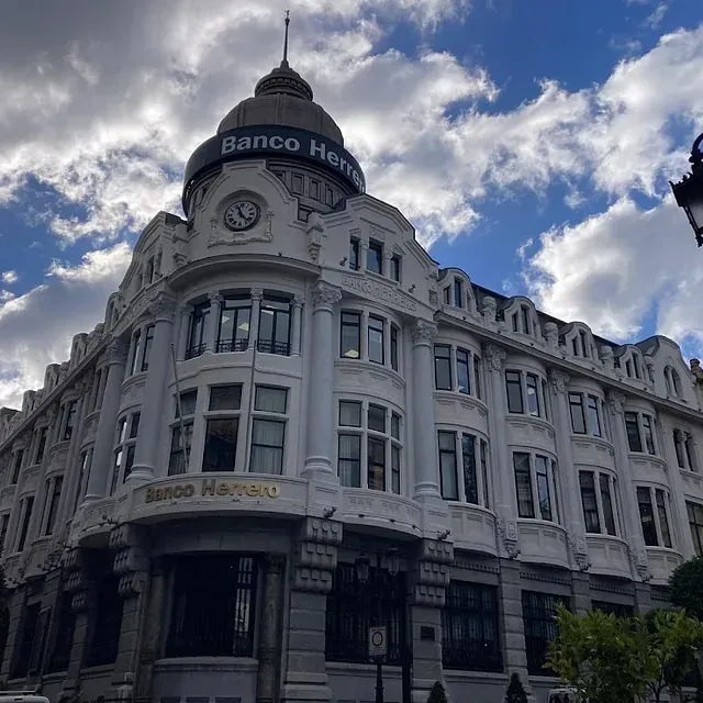 Edificio del Banco Herrero en Oviedo, España, con una torre de reloj, contra un cielo azul con nubes.