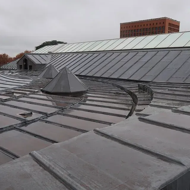 Techo metálico con secciones curvas y pequeñas aberturas cónicas. Al fondo se ve un edificio de ladrillo.