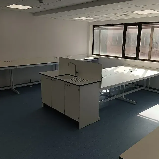 Laboratorio vacío y luminoso con mesas blancas, lavabo y suelo azul. Las ventanas dejan entrar la luz del sol.