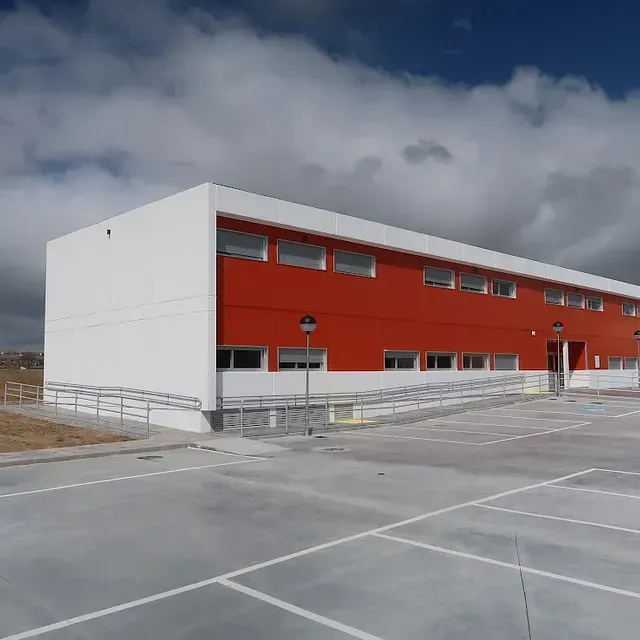 Edificio moderno con fachada naranja y blanca, al frente cuenta con rampa y amplio estacionamiento vacío.