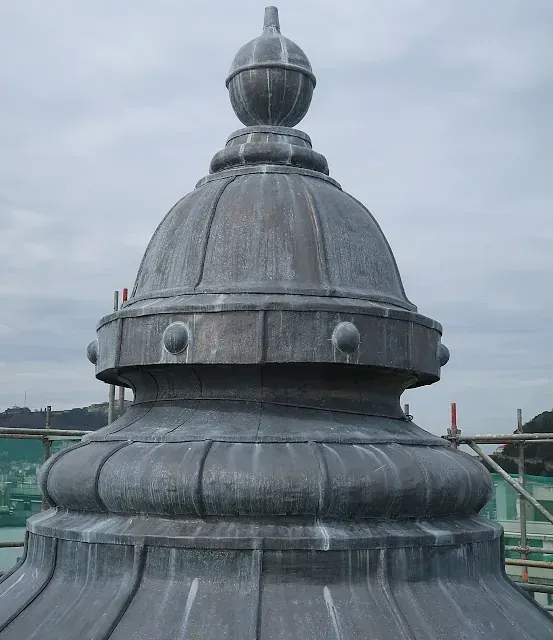 Cúpula con un remate decorativo en un edificio, gris contra un cielo nublado.