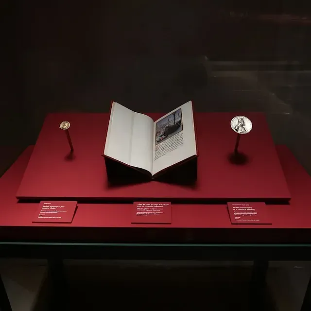 Una vitrina con un libro abierto, dos medallas y placas informativas sobre una superficie roja.