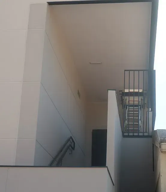 Vista exterior de un edificio con escaleras, barandillas metálicas y una silla en un pequeño rellano. Paredes beige claro.
