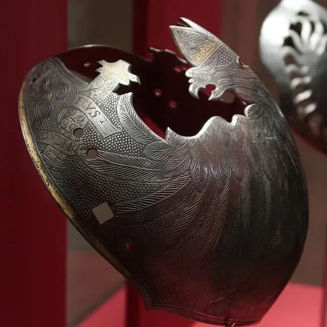 Fragmento de casco de acero dañado, con grabados ornamentales, posiblemente una armadura. Detalles: gris oscuro, patrones intrincados, pájaro estilizado.