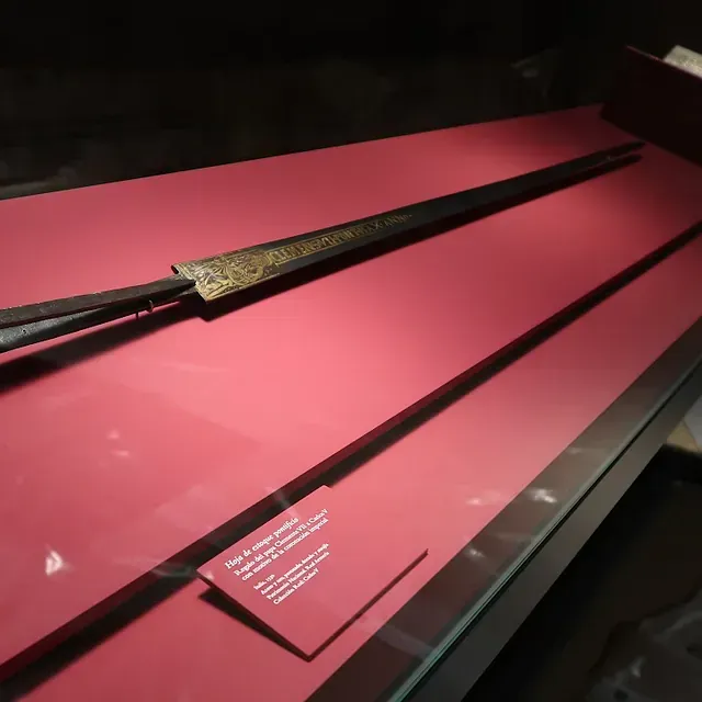 Espada larga y ornamentada en vitrina con fondo rojo y placa informativa.