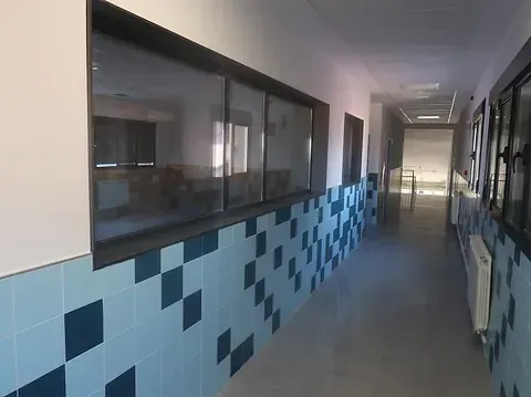 Pasillo largo en un edificio con paredes de azulejos azules y blancos, ventanas e iluminación brillante.