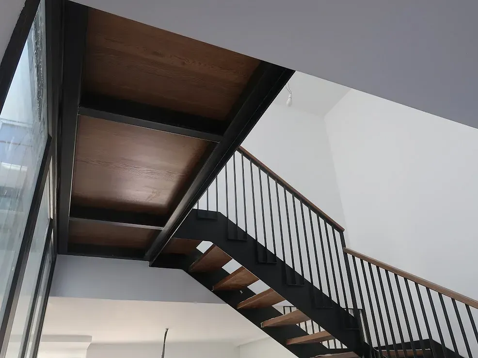 Escalera con estructura de metal negro, peldaños de madera y barandilla. Interior con paredes blancas y una ventana.