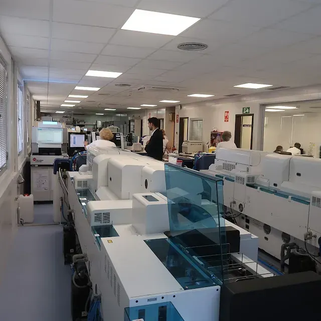 Laboratorio con equipo de pruebas automatizado; personal trabajando. Iluminación fluorescente. Máquinas blancas y grises.