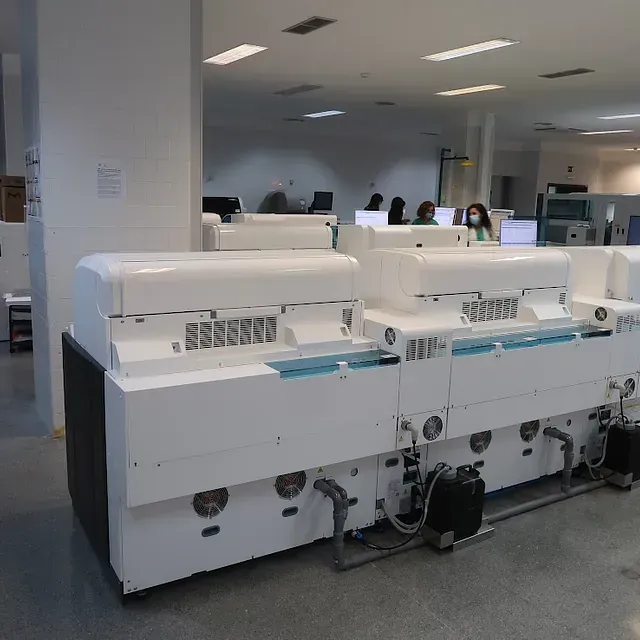 Equipo de laboratorio: grandes analizadores blancos en un laboratorio moderno, con gente en el fondo.