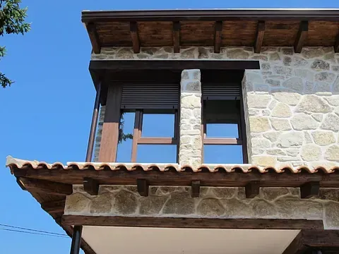Casa de piedra con marcos de ventanas de madera, contraventanas y techo de tejas bajo un cielo azul.