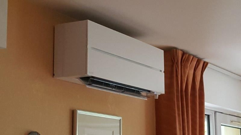 Unité de climatisation murale blanche sur un mur beige, à côté d'un rideau orange.