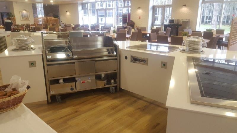 Espace buffet avec appareils électroménagers en acier inoxydable et comptoirs blancs ; salle à manger en arrière-plan.