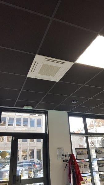 Unité de climatisation fixée au plafond dans une pièce au plafond noir à caissons. Fenêtres et bâtiment visibles.