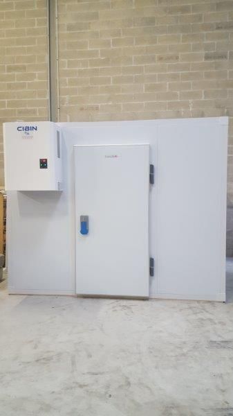 Chambre froide industrielle blanche adossée à un mur de briques, avec porte et panneau de commande intégré.