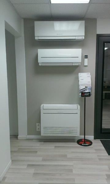 Trois unités de climatisation blanches sont exposées sur un mur gris dans une salle d'exposition.