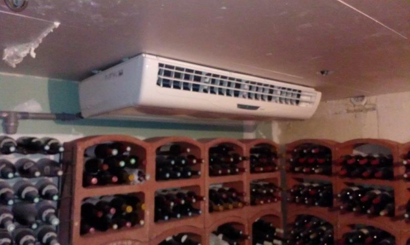 Cave à vin avec rangées de bouteilles sur étagères, unité de réfrigération fixée au plafond.