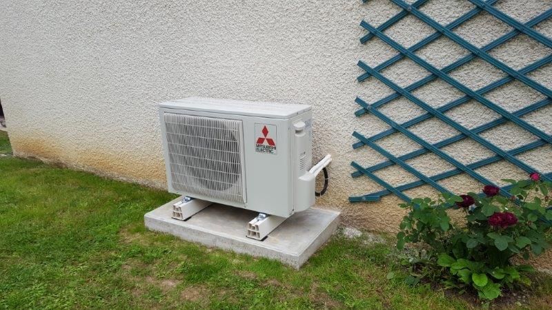 Unité de climatisation Mitsubishi sur une base en béton, à côté d'un mur et d'une treille.