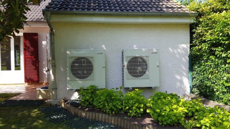 Deux unités de climatisation extérieures fixées sur un mur blanc d'une maison, au-dessus de buissons verts.